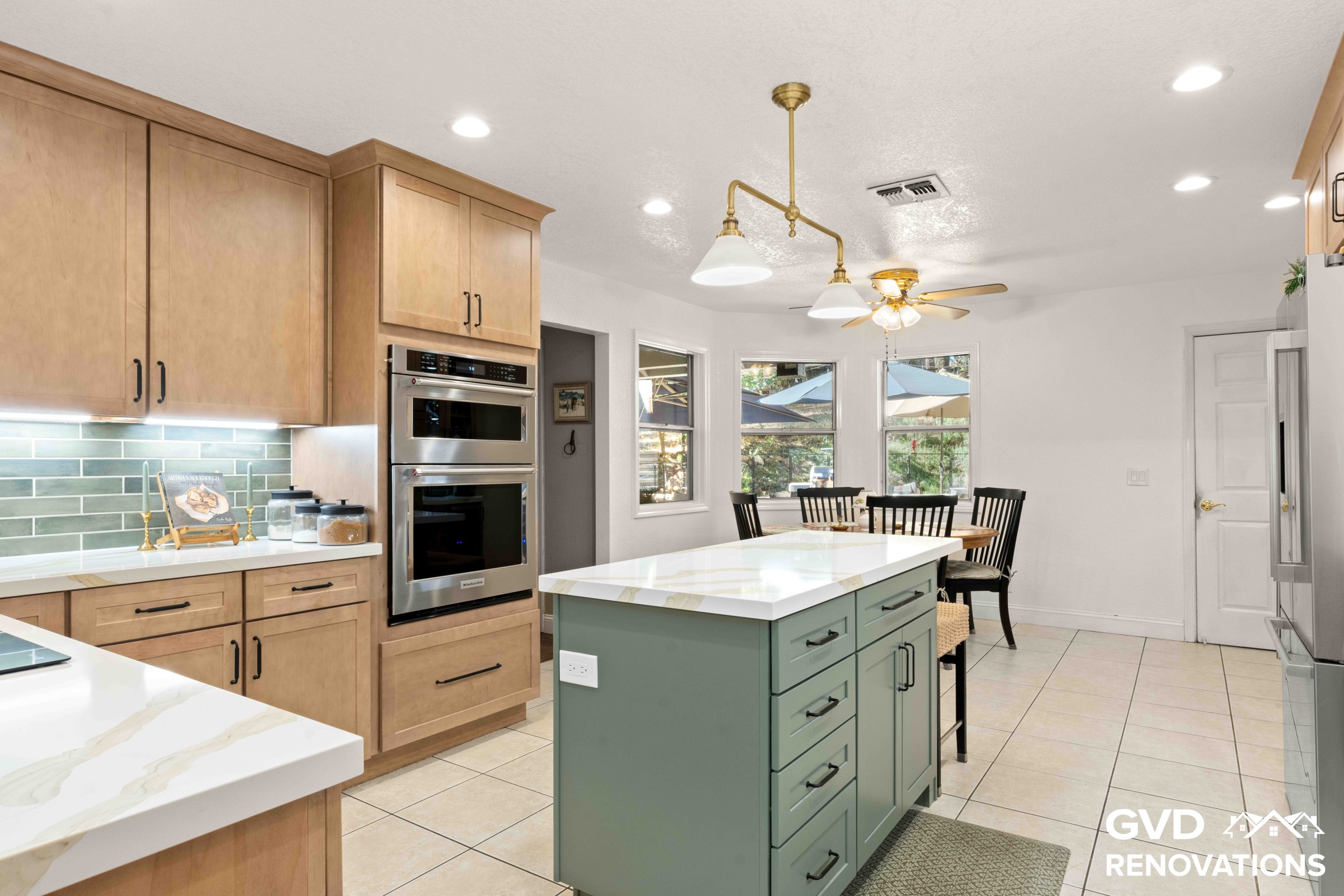 Open kitchen with a sage green island, schoolhouse pendant lights, stainless wall oven and an adjacent dining nook