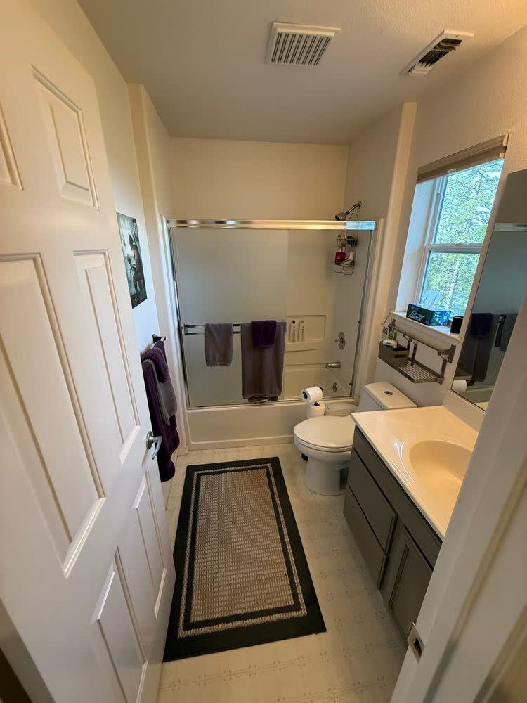 Before: original hall bathroom with a tub-and-shower combo behind sliding glass, a grey vanity and a forest-view window
