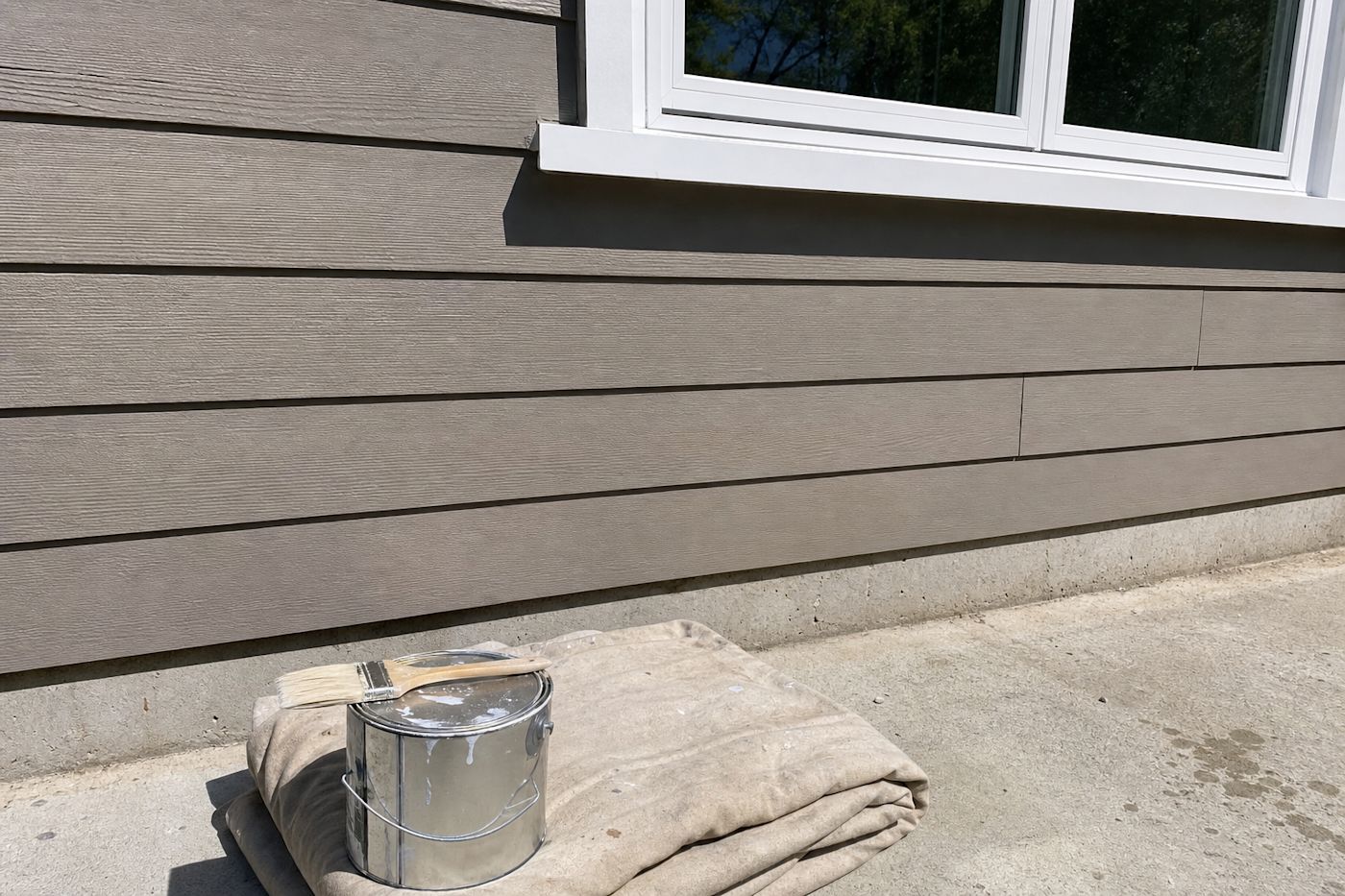 A paint can, brush and drop cloth beside an engineered wood lap siding wall, showing the repaint and maintenance cycle