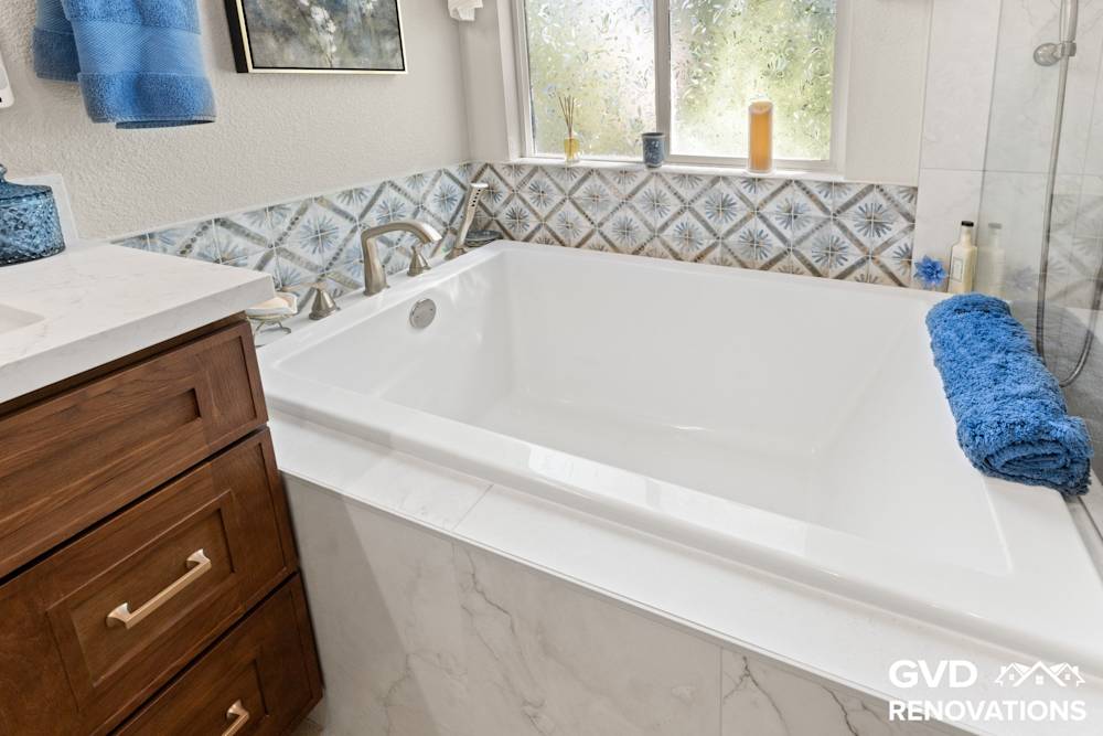 Drop-in soaking tub beneath a window with a marble tile deck and blue accent tile