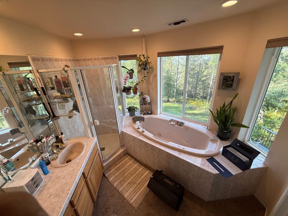 Before: original primary bathroom with an oak double vanity, a corner glass shower stall and an oval drop-in jetted tub beneath the forest-view windows