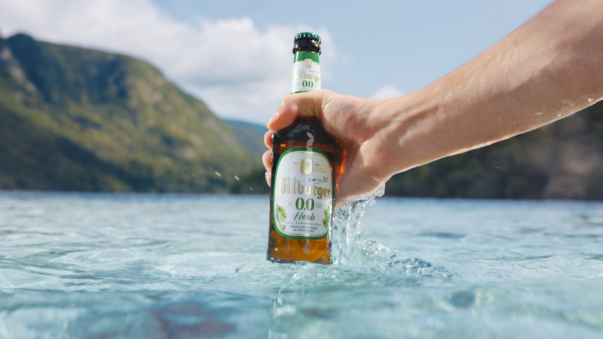Eine Flasche Bitburger 0,0 Herb wird mit einer Hand aus einem Bergsee geholt