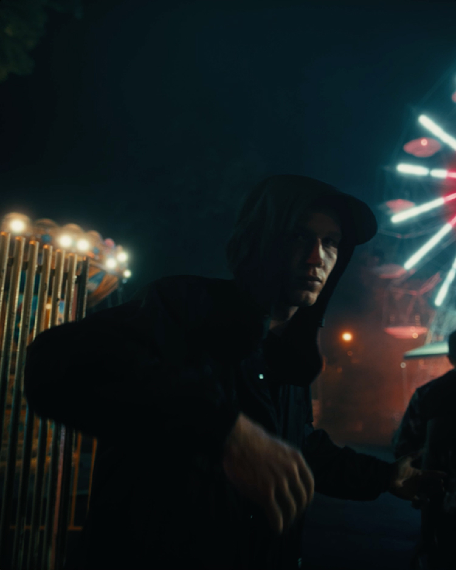 Vor einem schwarzen Hintergrund steht eine dunkel gekleidete Person mit Hut. Lichteffekte sind rechts und links sichtbar.