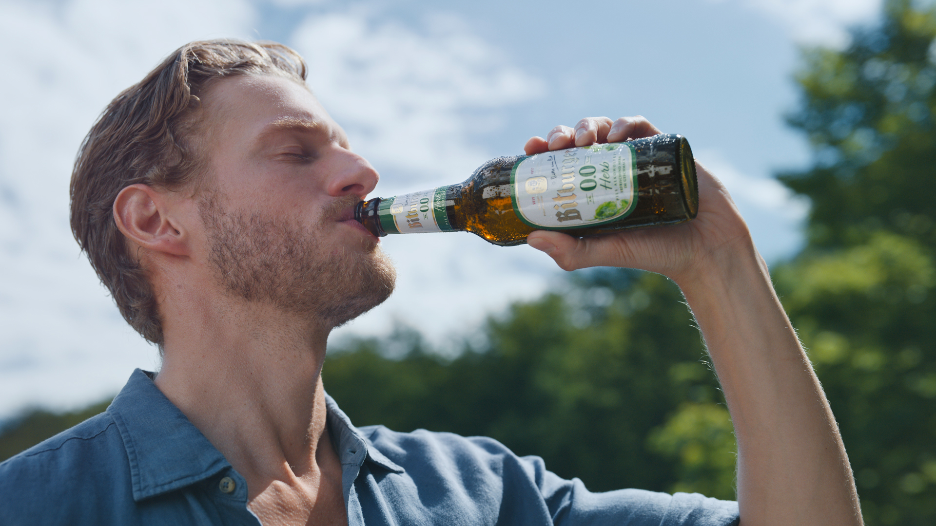 Ein junger Mann mit Bart steht in der Natur vor grünen Bäumen und trinkt ein Bitburger 0,0 Herb aus der Flasche