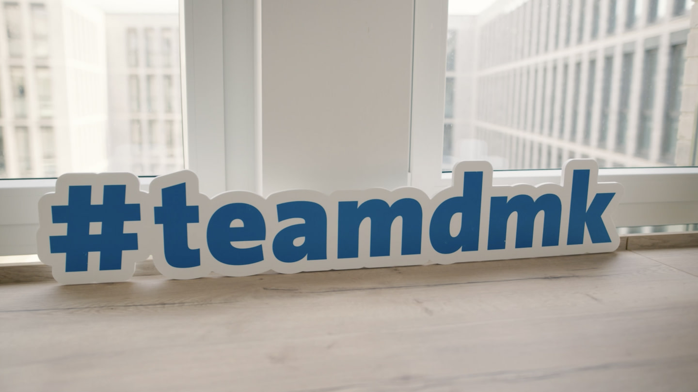 Case | DMK | Video Fallback "#teamdmk“ Schriftzug in Blau mit weißen Rand steht ausgeschnitten auf einer Fensterbank
