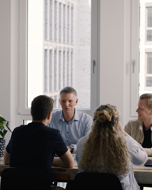 Case | DMK | Gallery 2 | Bild 2 Vier Menschen sitzen sich gegenüber an einem Tisch und reden