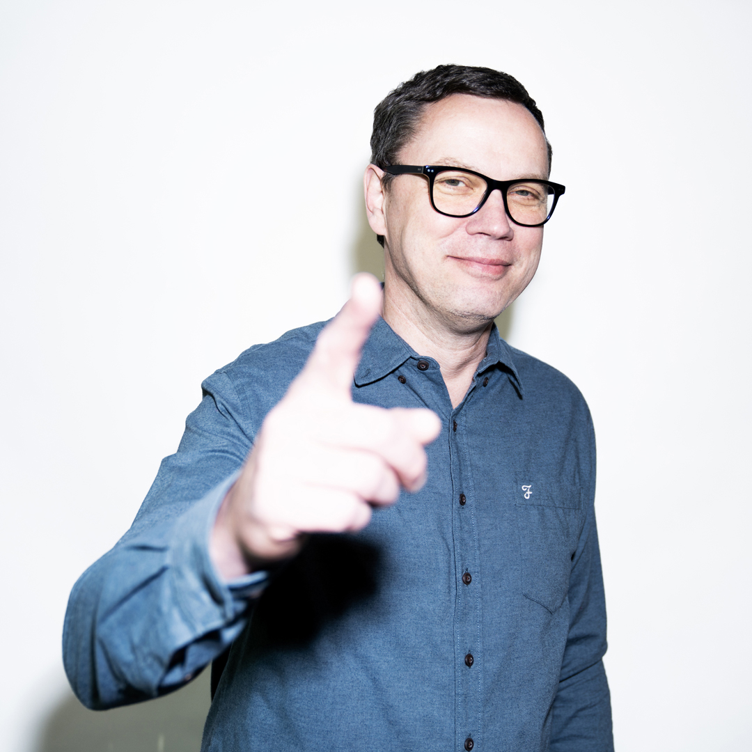 Heiko Quant steht in der Mitte, lächelt und zeigt in die Kamera