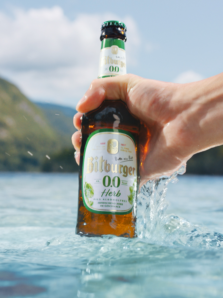 Eine Flasche Bitburger 0,0 Herb wird mit einer Hand aus einem Bergsee geholt
