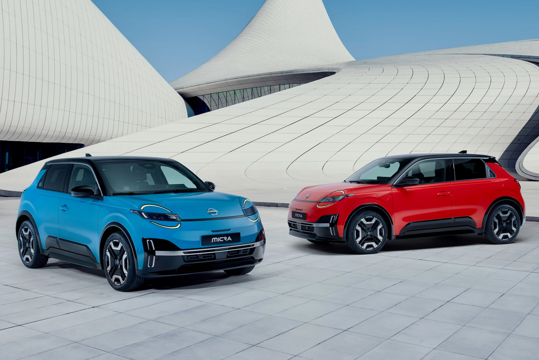 two iconic Micras - gear blue color and red color - parked in front of a blue & white design architectural building in a V shape facing each other