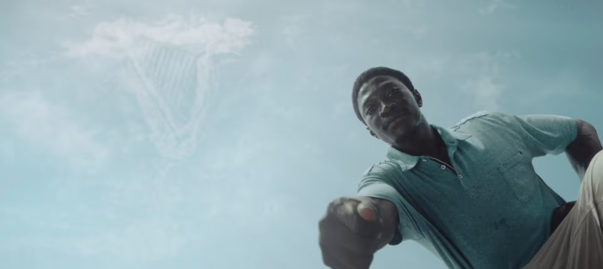 Looking up at the sky with clouds forming the shape of the Guinness logo. A person leans over the camera, holding a seed they are about to plant.
