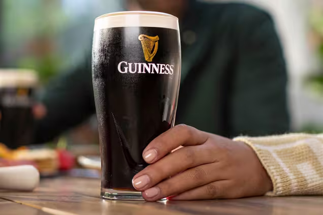 A full pint of Guinness is centre of the image, a hand resting on the table grips the base of the glass.