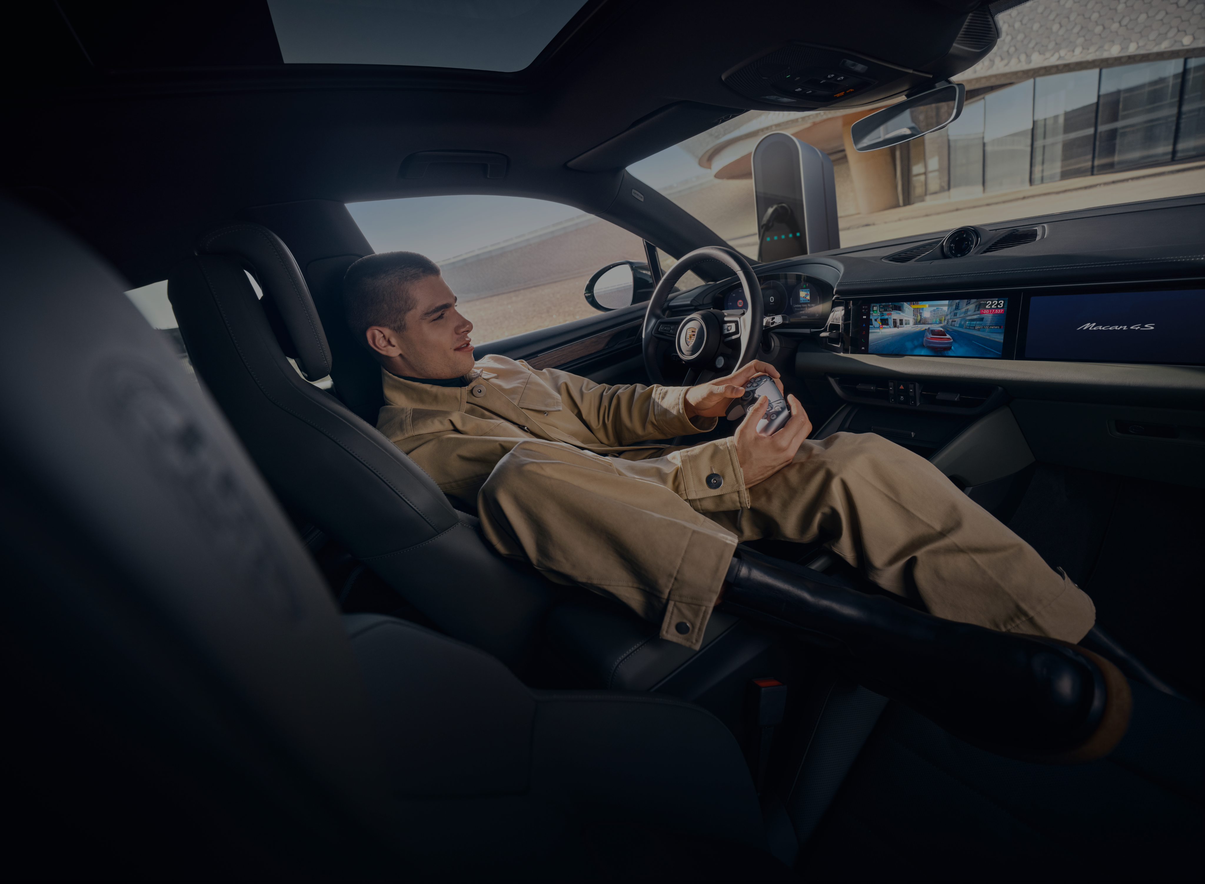 Person reclining in the driver’s seat of a modern car, holding a gaming controller and playing a racing game displayed on the dashboard screen.