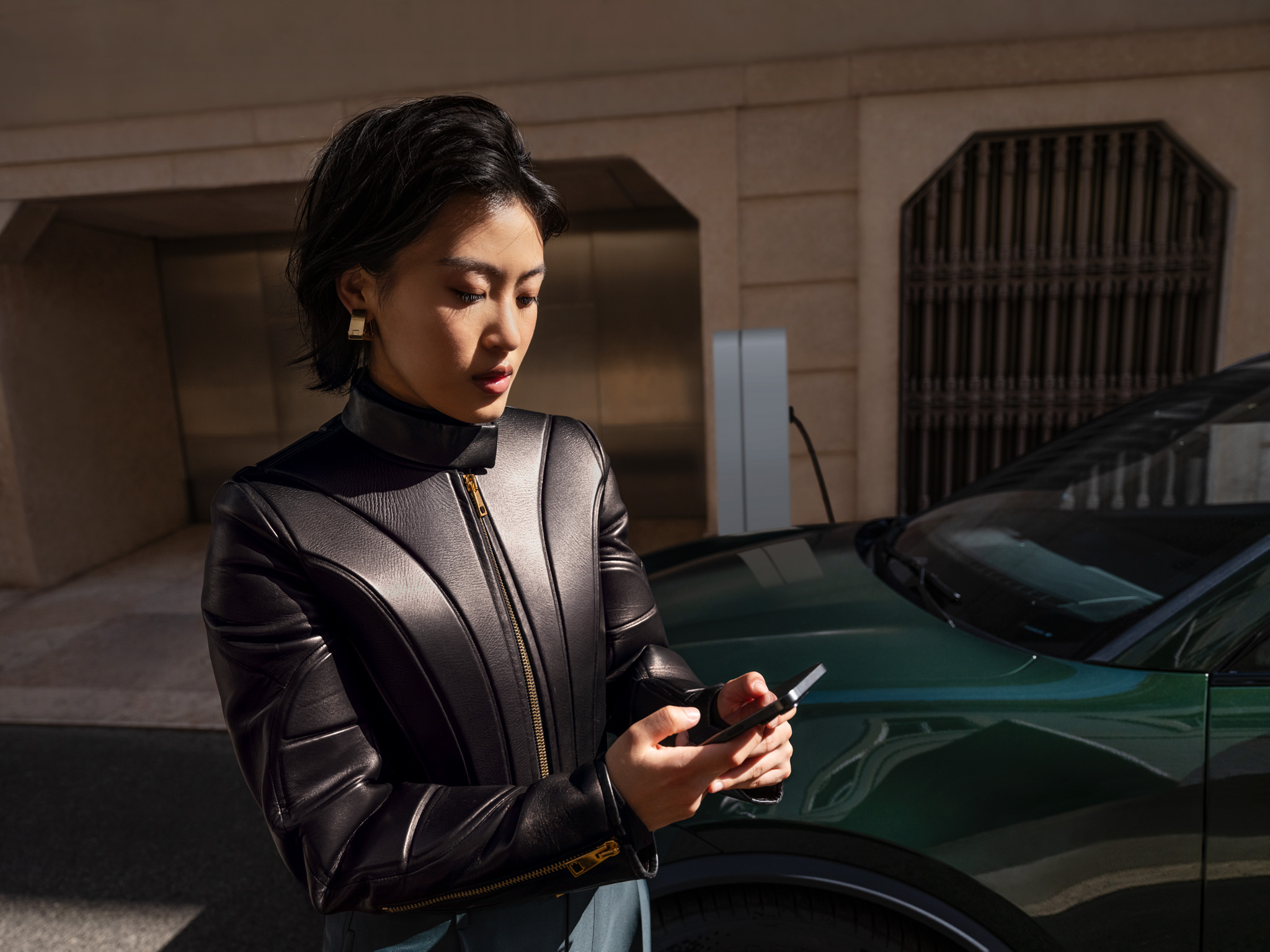 A stylish woman in a black leather jacket stands next to a dark green electric vehicle, using her smartphone. The vehicle is connected to a charging station in a modern urban setting. Sunlight casts sharp shadows, emphasizing the clean lines of the architecture and the sleek design of the car.