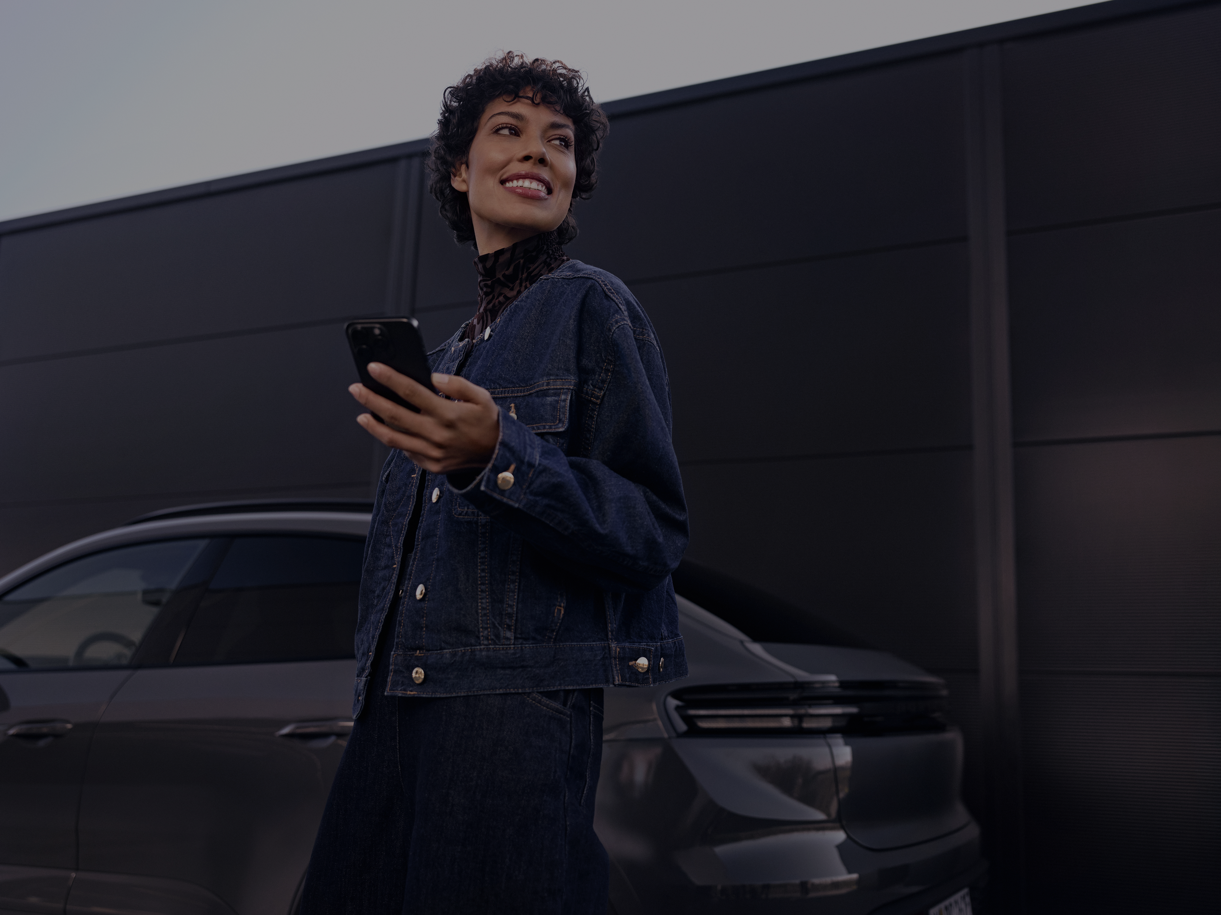 A smiling person wearing a denim jacket stands outdoors beside a modern car while holding a smartphone and looking into the distance.