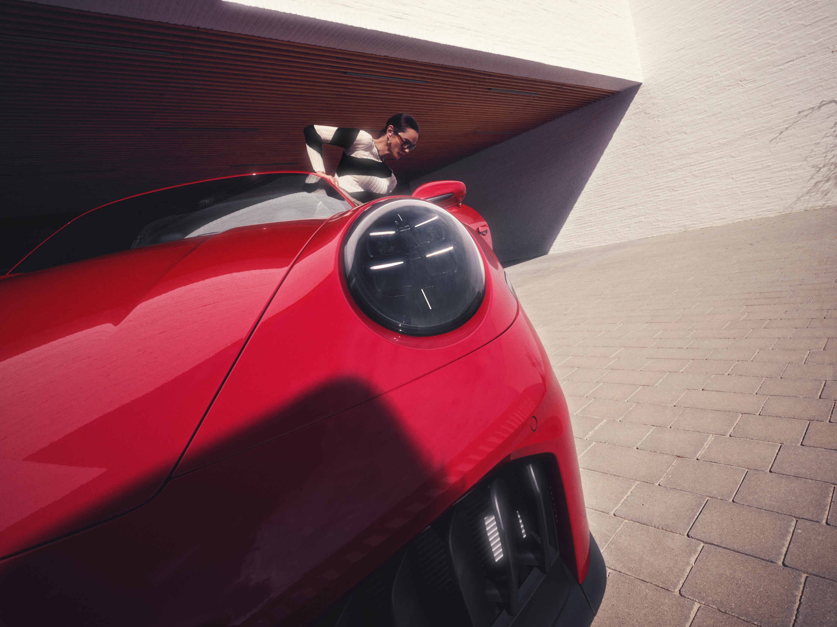 A woman in a black-and-white outfit leans over a sleek, red sports car parked in front of a modern architectural structure with sharp angles and white brick walls. The image is captured from a dramatic low angle, emphasizing the car’s front headlight and aerodynamic curves.