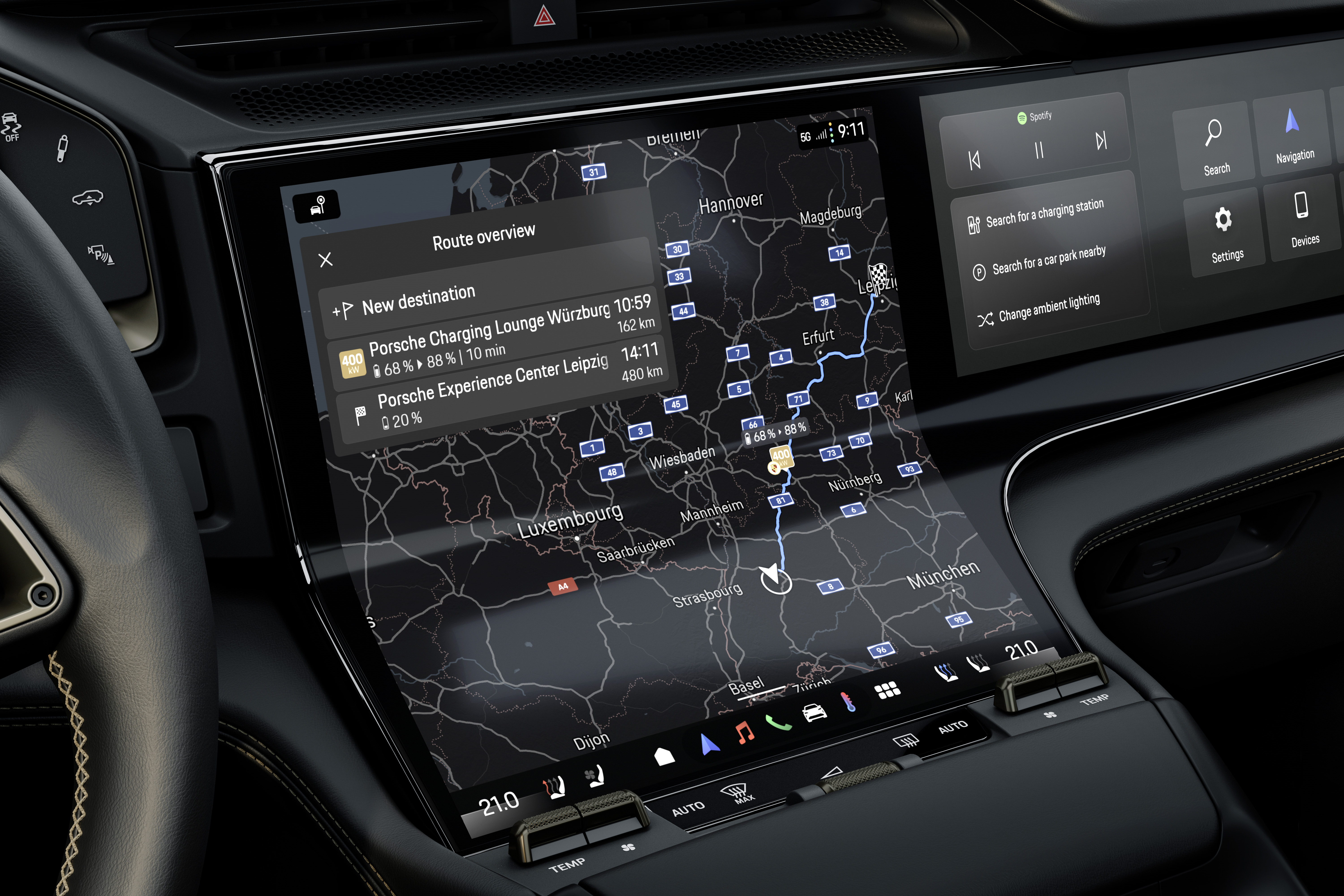 Inside a modern Porsche, a woman drives through a city street lined with palm trees and skyscrapers. A man in the passenger seat, wearing headphones and a smartwatch, interacts with the touchscreen display in front of him, playing a racing video game. The dashboard shows navigation directions, and a Porsche-branded travel mug sits in the center console. The woman's face is reflected in the rearview mirror.