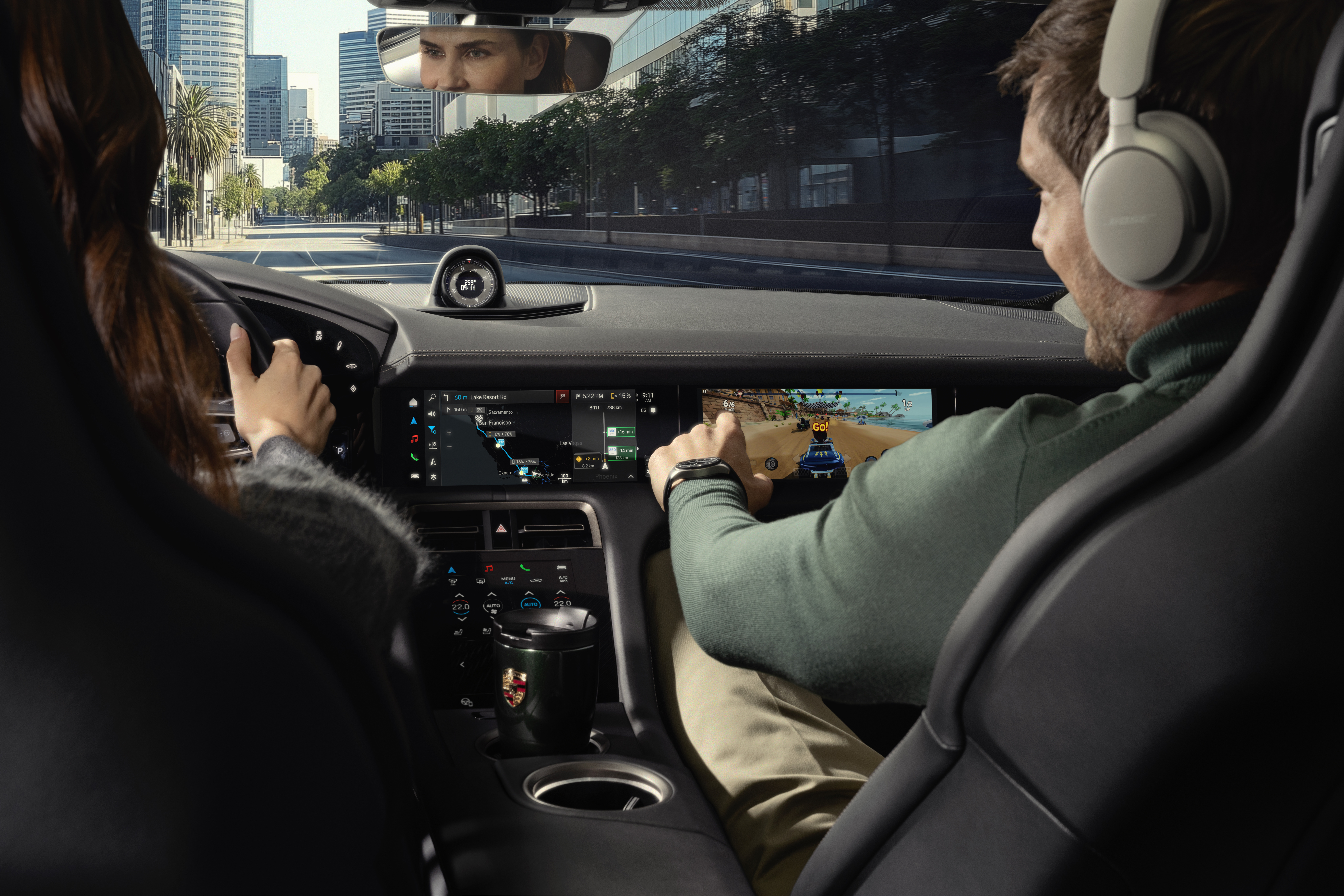 Inside a modern Porsche, a woman drives through a city street lined with palm trees and skyscrapers. A man in the passenger seat, wearing headphones and a smartwatch, interacts with the touchscreen display in front of him, playing a racing video game. The dashboard shows navigation directions, and a Porsche-branded travel mug sits in the center console. The woman's face is reflected in the rearview mirror.