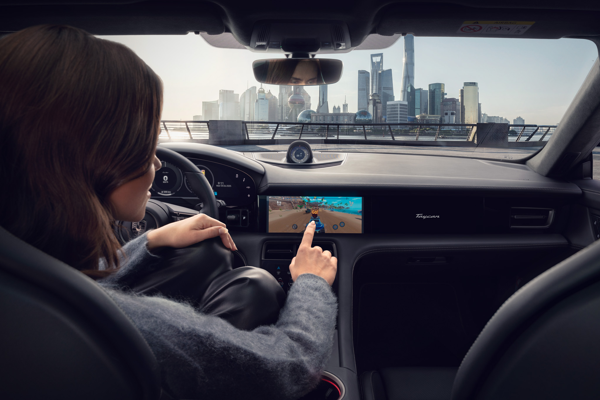 A person seated in the driver’s seat of a car interacts with a touchscreen display showing a racing game, with a modern city skyline visible through the windshield in the background.