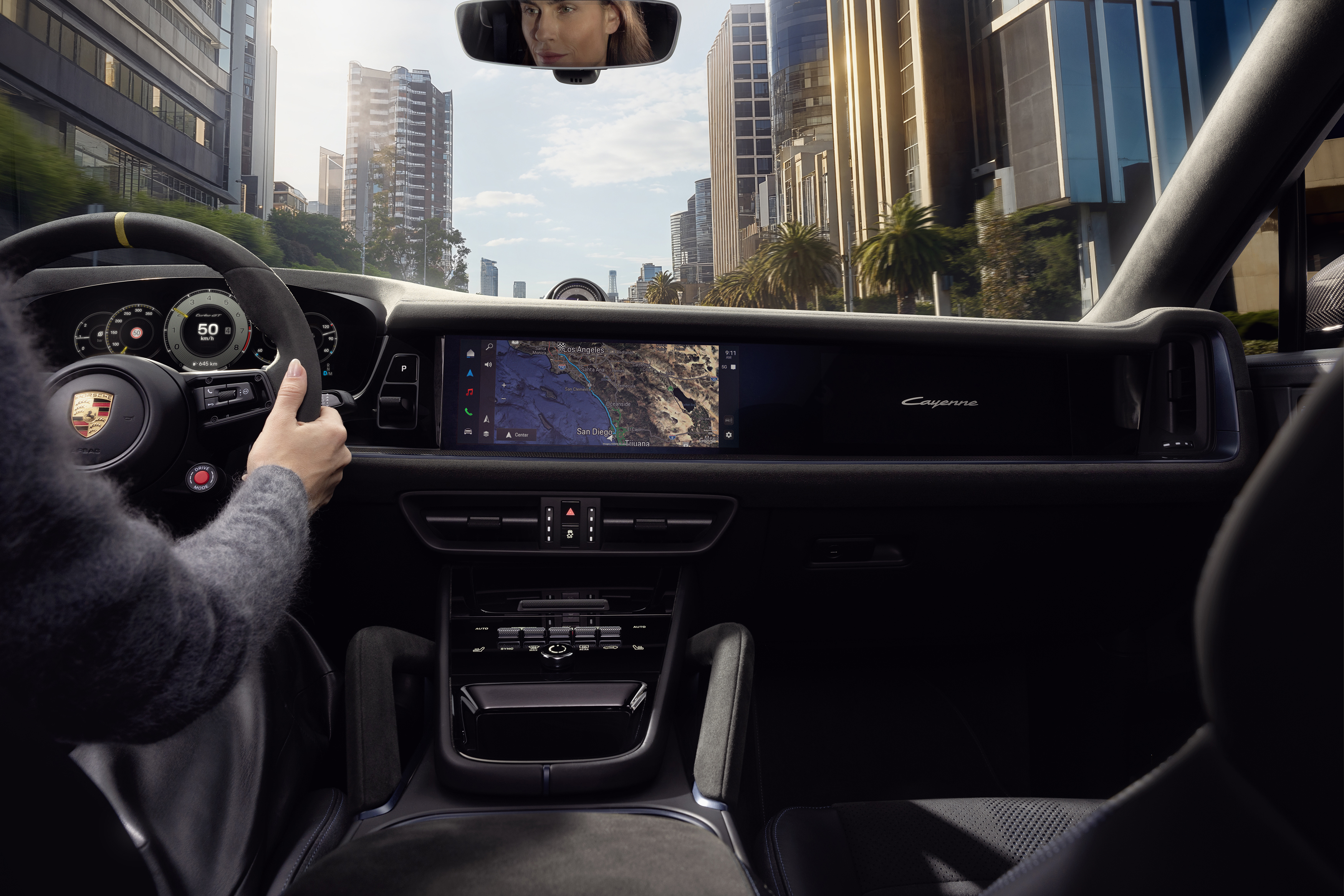 Interior view of a Porsche Cayenne being driven through a city with tall, modern buildings and palm trees. The driver grips the steering wheel, and the dashboard displays a digital speedometer reading 50 mph and a large central screen showing a satellite navigation map of Southern California. The passenger-side screen displays the “Cayenne” logo, and the driver’s reflection is visible in the rearview mirror.