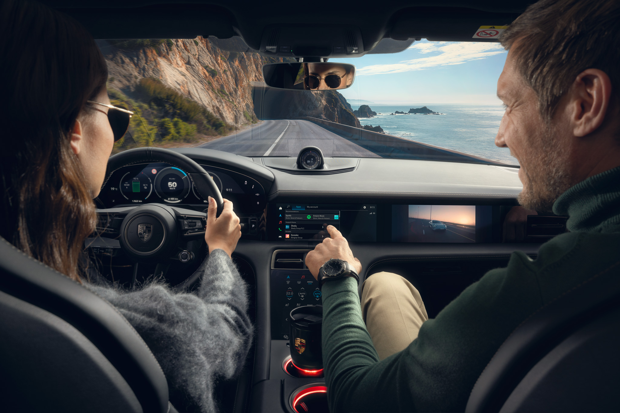 A man and woman are driving along a scenic coastal road in a Porsche. The woman is behind the wheel, wearing sunglasses, while the man in the passenger seat interacts with the central touchscreen. The dashboard displays navigation and media content, including a video of a car driving at sunset. The digital instrument cluster shows a speed of 50 km/h. The ocean and rugged cliffs are visible through the windshield, reflecting a peaceful, high-tech driving experience.