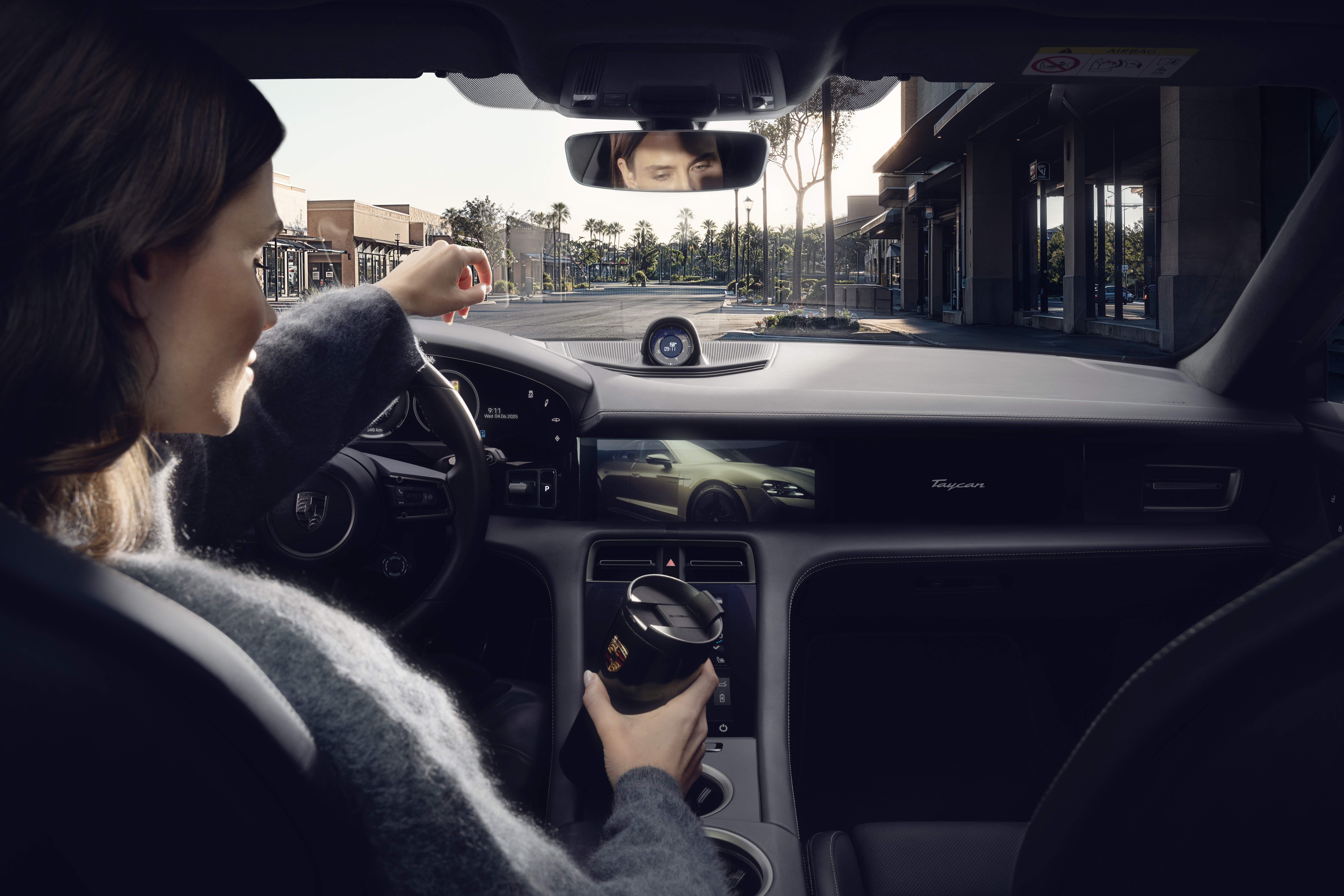 A woman sitting in a Porsche in an urban environment, watching a video on the display screen while holding a black coffee mug in her hand.