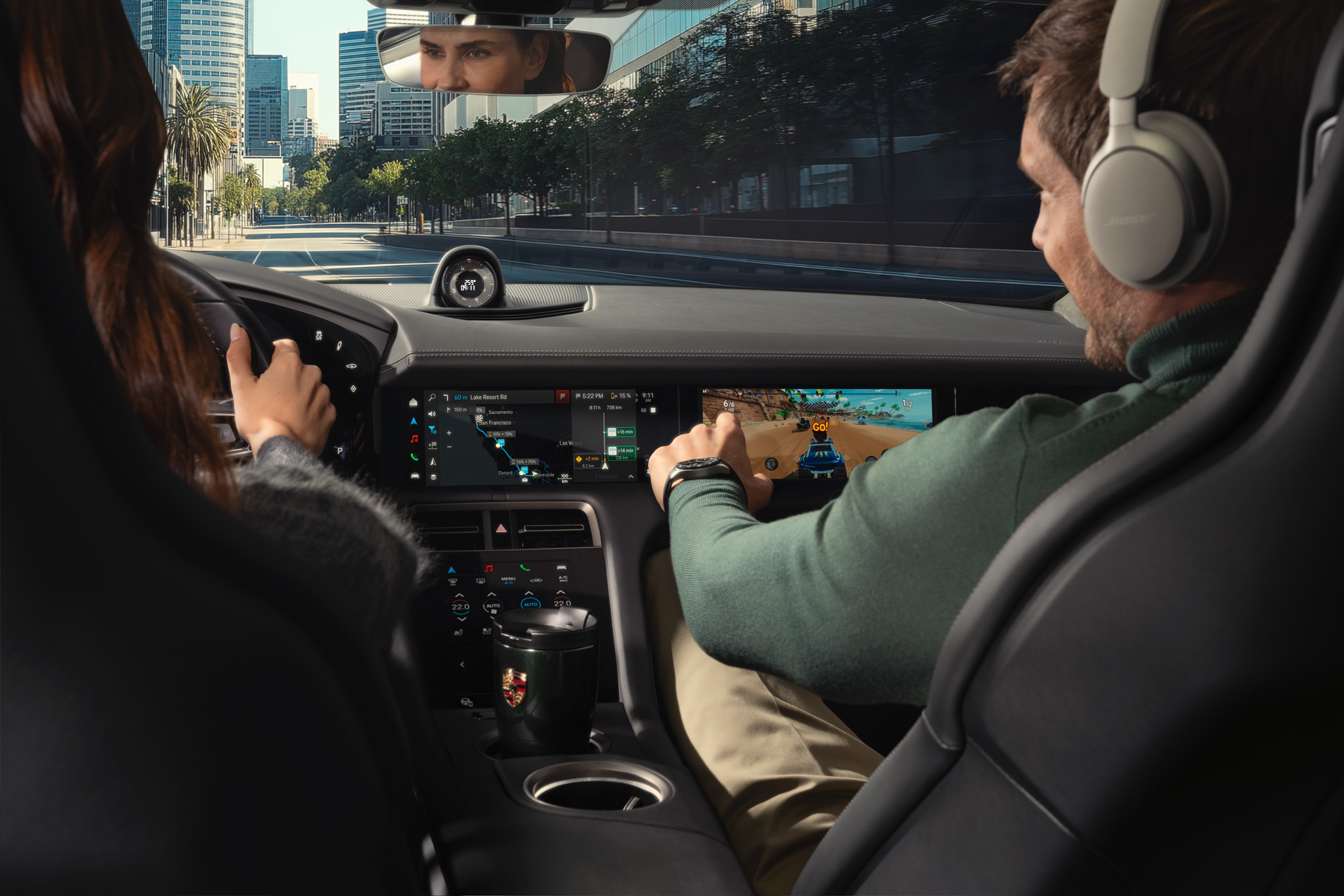 Interior view of a car with the driver focused on the road ahead, while the front passenger wearing headphones interacts with a touchscreen displaying a racing game; the center display shows navigation guidance through a city street.