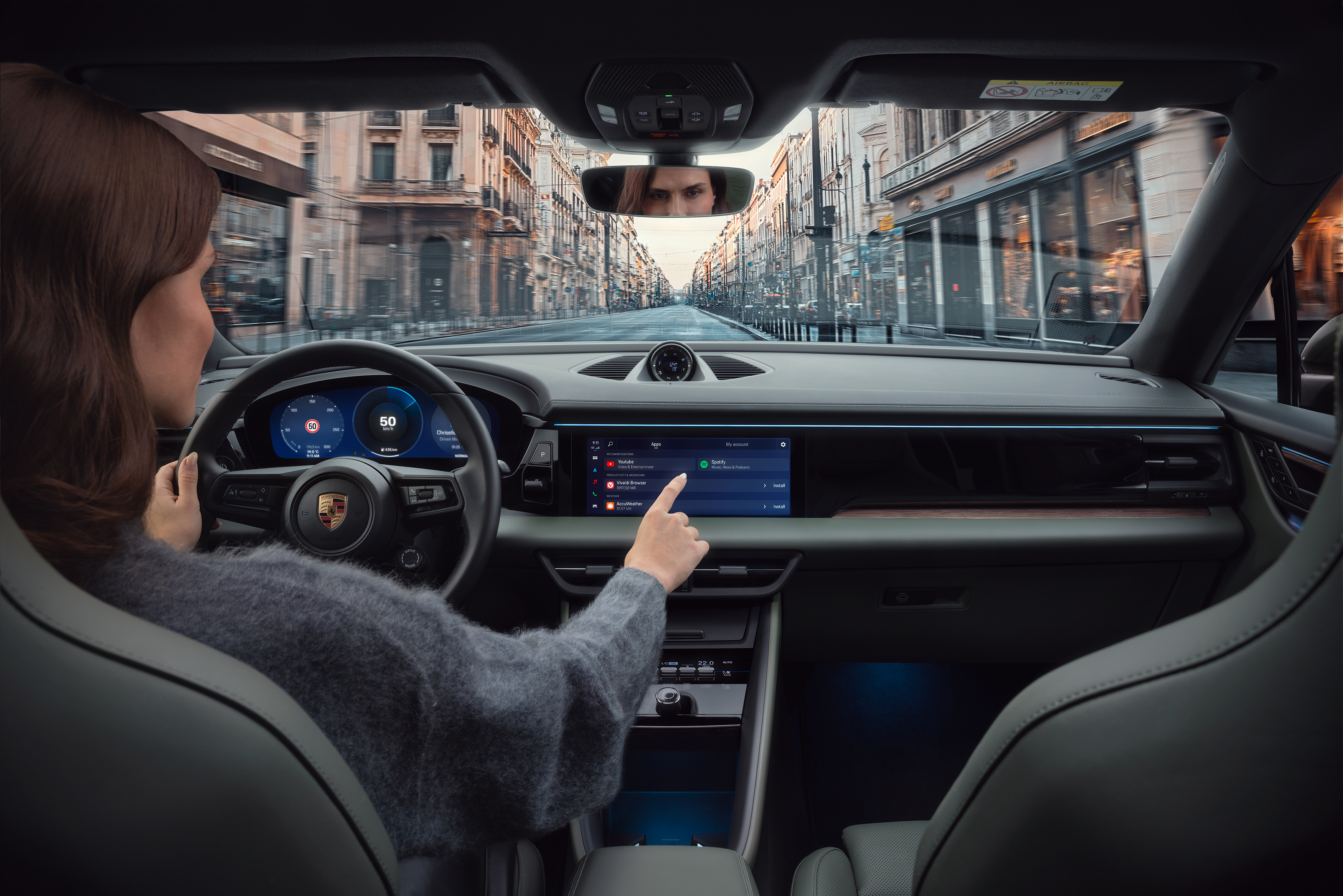 A woman sitting in a Porsche in an urban environment, touching the display of the car. The display displays some Apps such as music streaming and navigation programs.