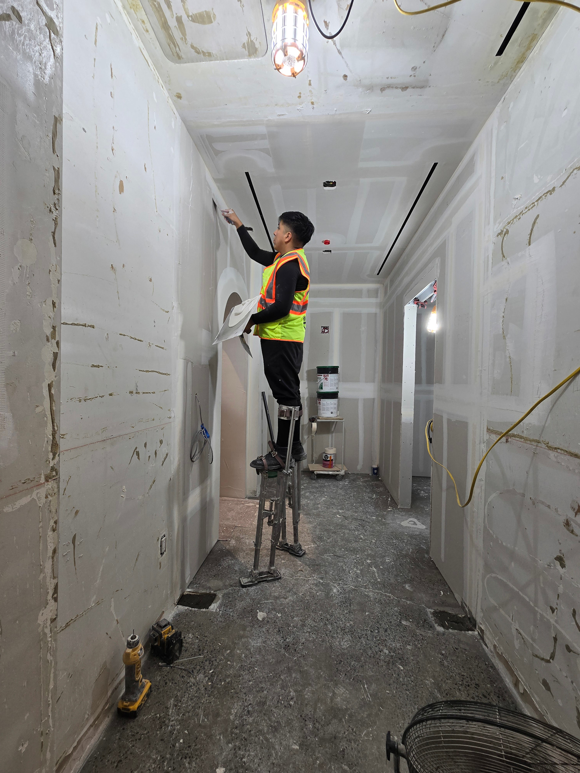Worker in safety vest on ladder inspecting drywall ceiling in construction hallway.