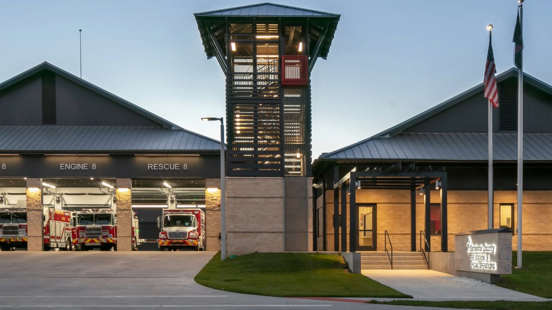 Modern fire station exterior at dusk with illuminated training tower, engine bays housing red fire trucks, and American flag