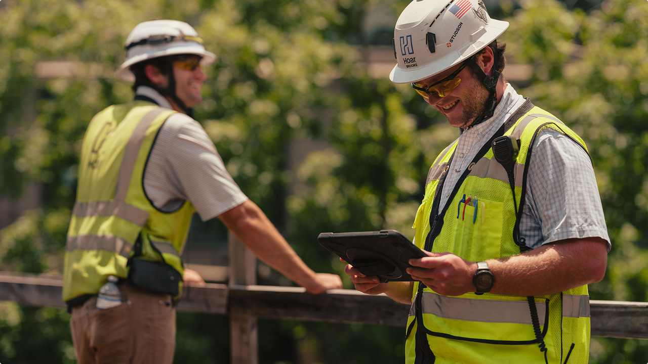 Two construction workers in yellow vests and helmets, one using a tablet.