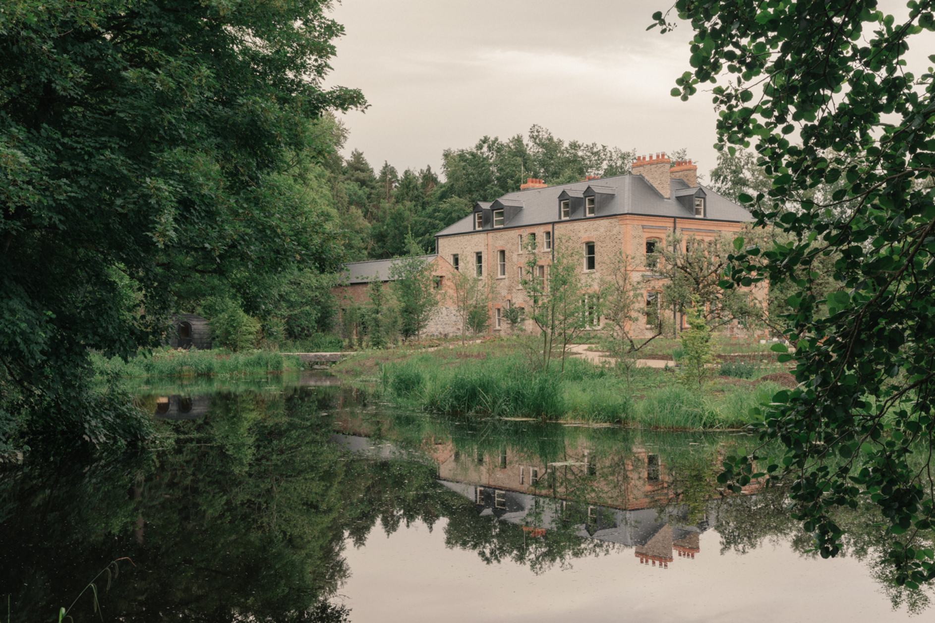 Brick mansion reflected in calm pond, surrounded by lush green trees and vegetation.