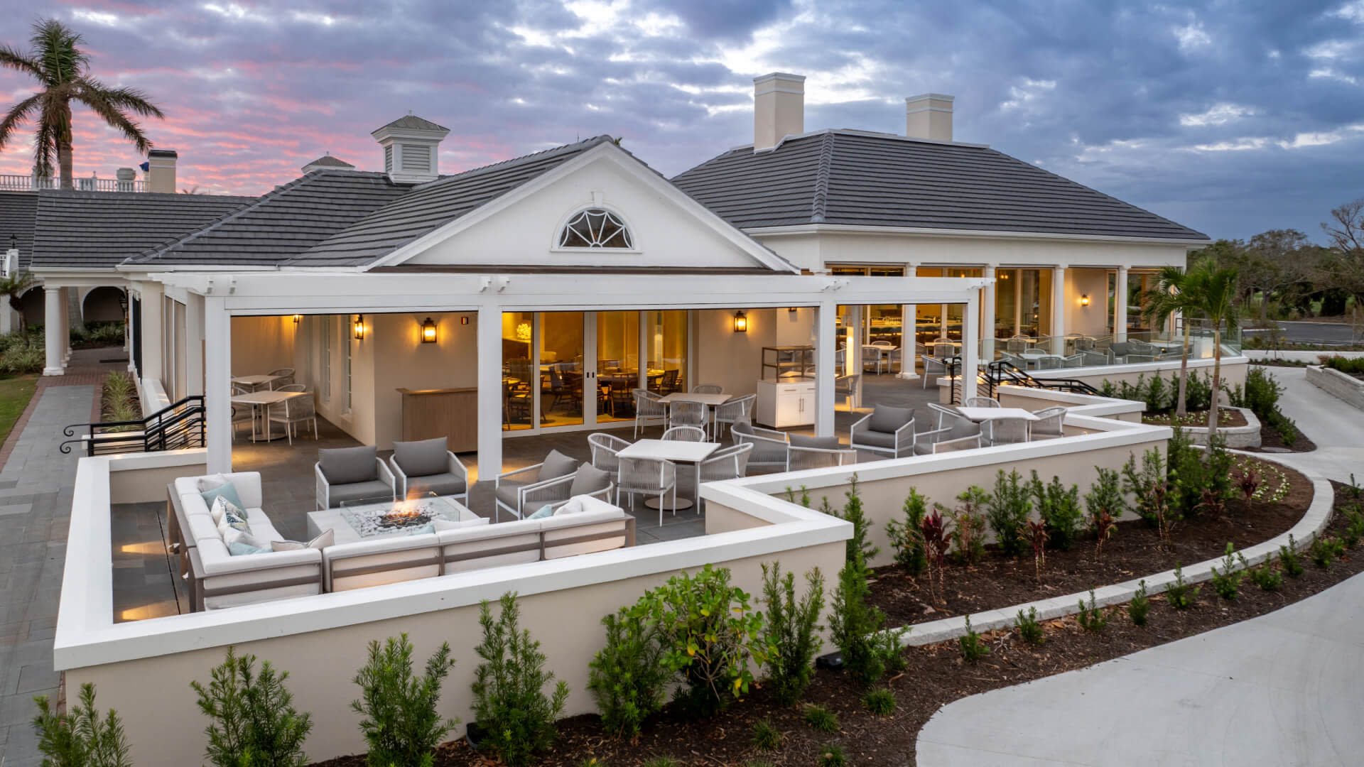 Elegant white clubhouse with outdoor patio featuring modern furniture, fire pit, and landscaped gardens at dusk under dramatic sky