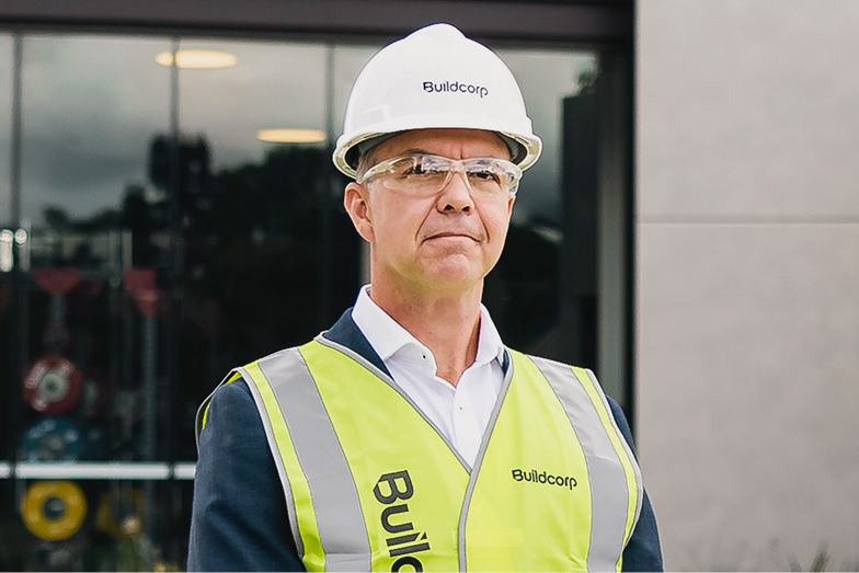 Man in white hard hat and yellow safety vest standing outside a building.