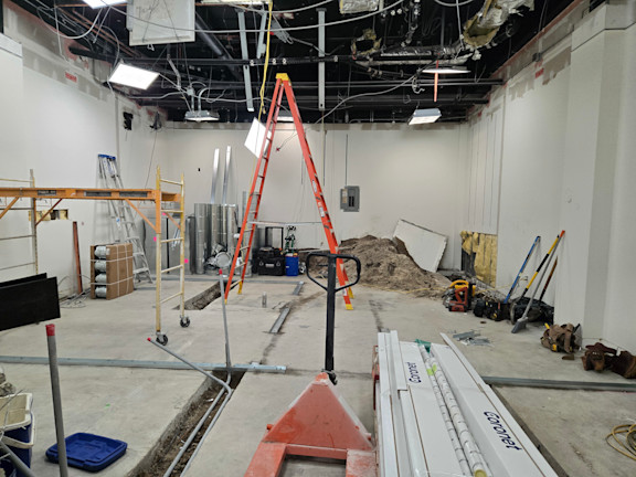 Construction site interior with red ladder, exposed ceiling, building materials, and equipment scattered throughout unfinished space.