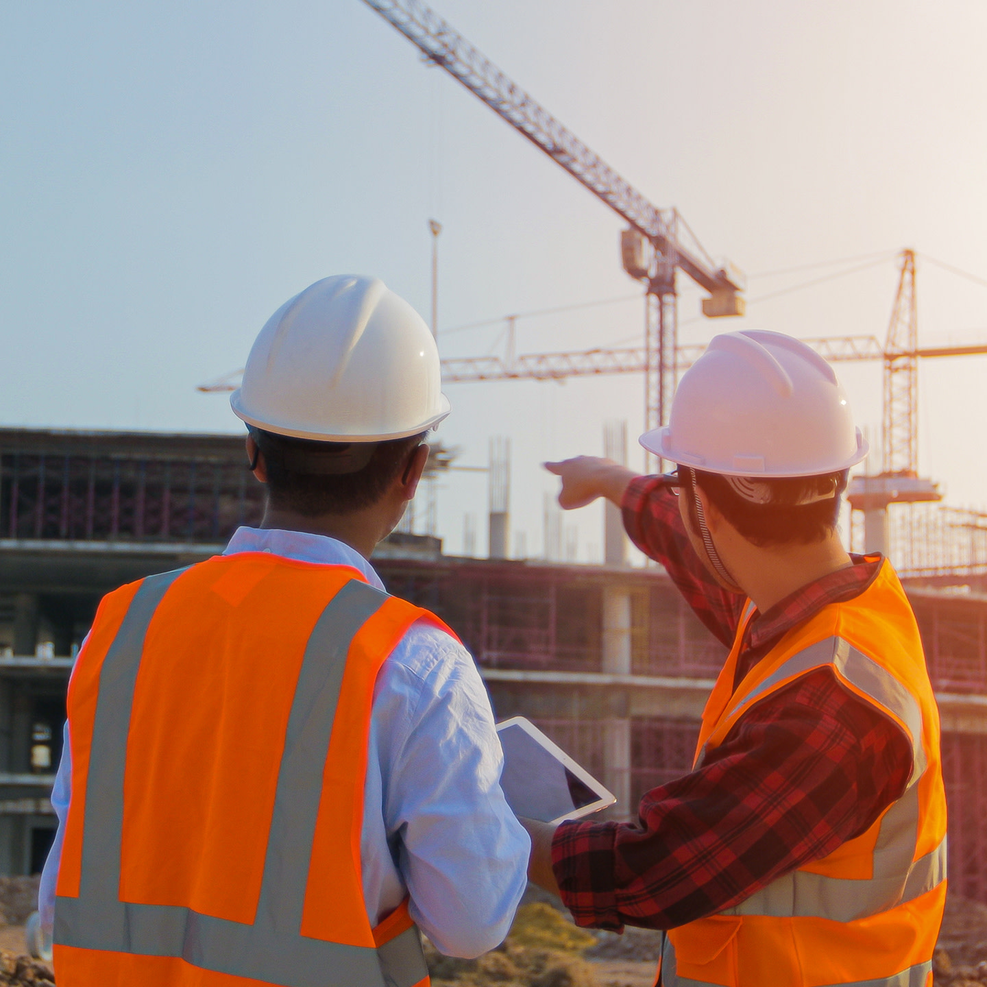 Two construction workers holding a tablet while pointing to a building under construction