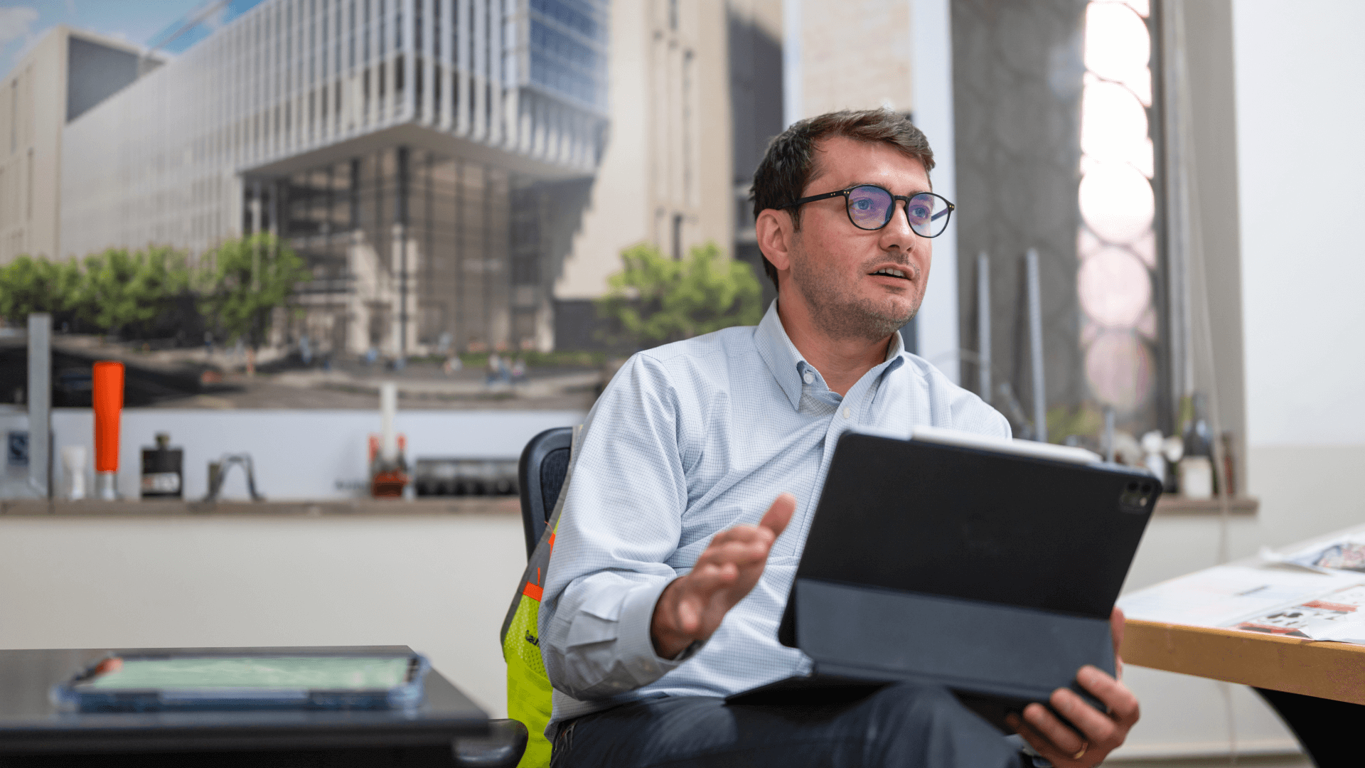 Man in glasses holding a tablet, gesturing while seated indoors.