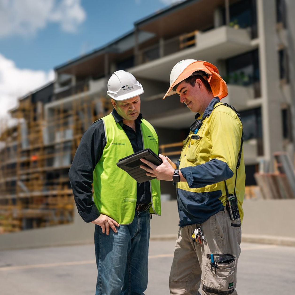 Two contractors wearing safety vests, standing at a construction site while using a tablet