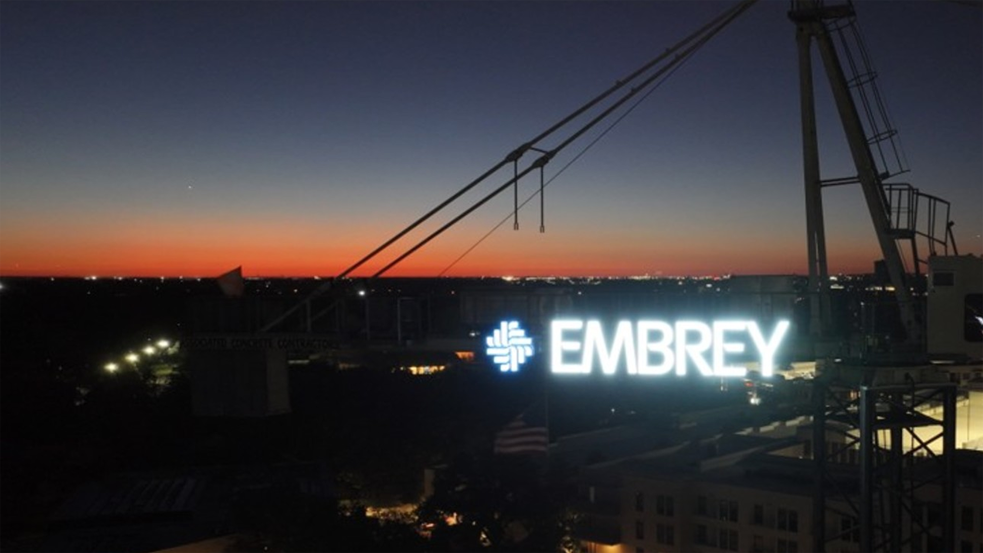 Illuminated EMBREY sign at dusk with suspension bridge and orange sunset sky
