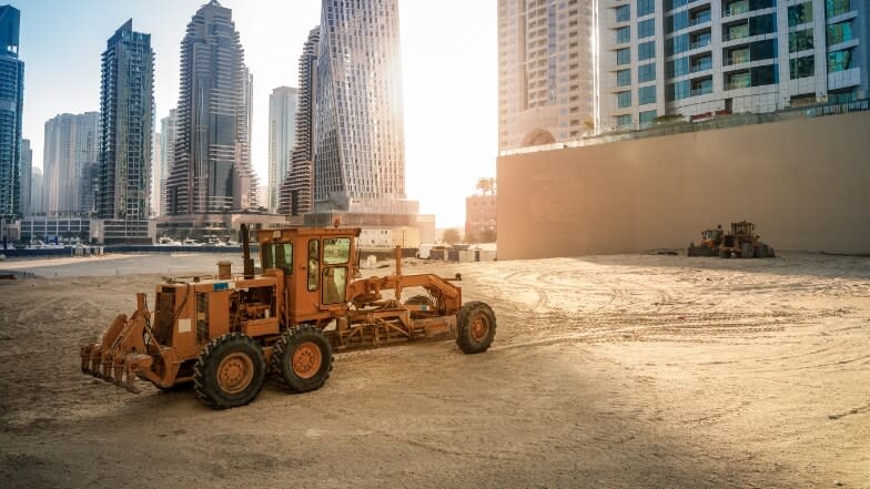 A grader machine parked in the middle of an empty construction site with skyscrapers on the back