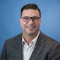 Professional headshot of smiling man wearing glasses and gray blazer against blue background.