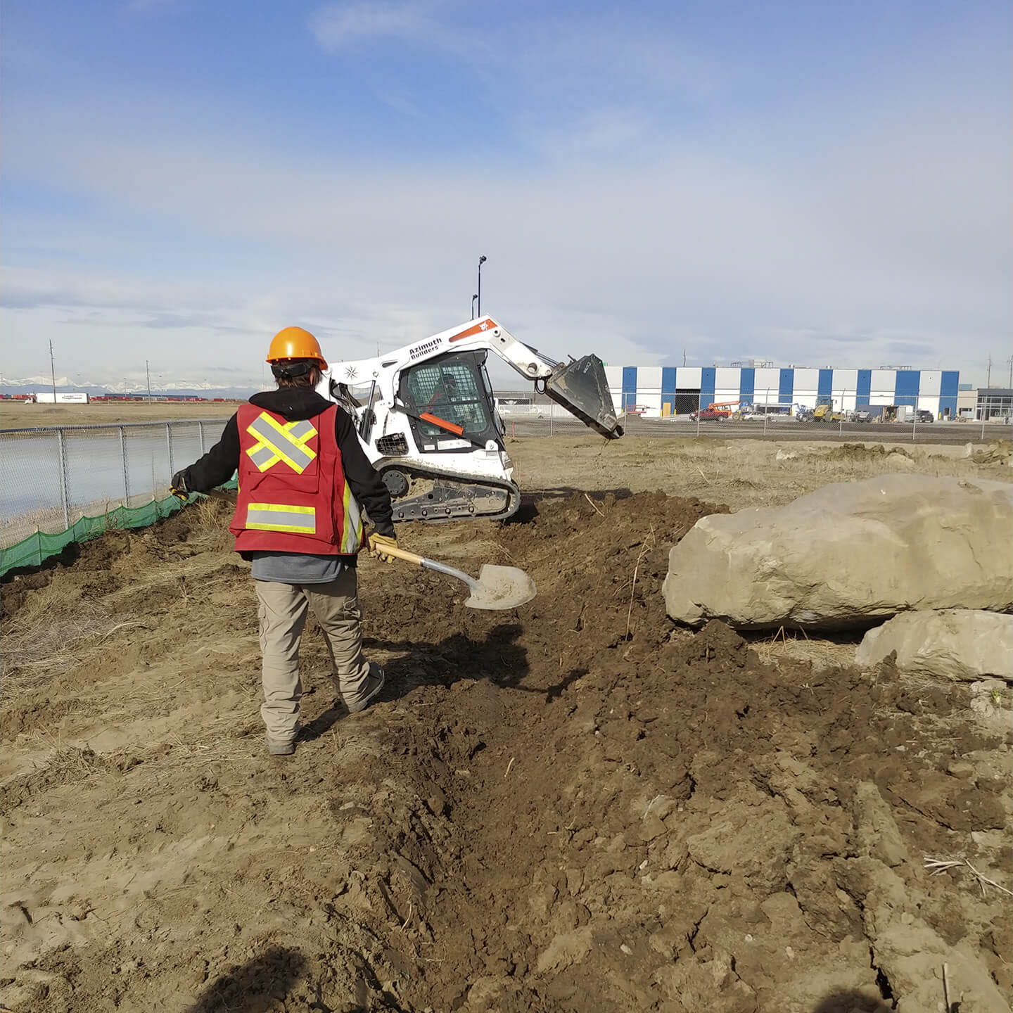 Azimuth Builder worker walking with a shovel and an excavator digging in the back