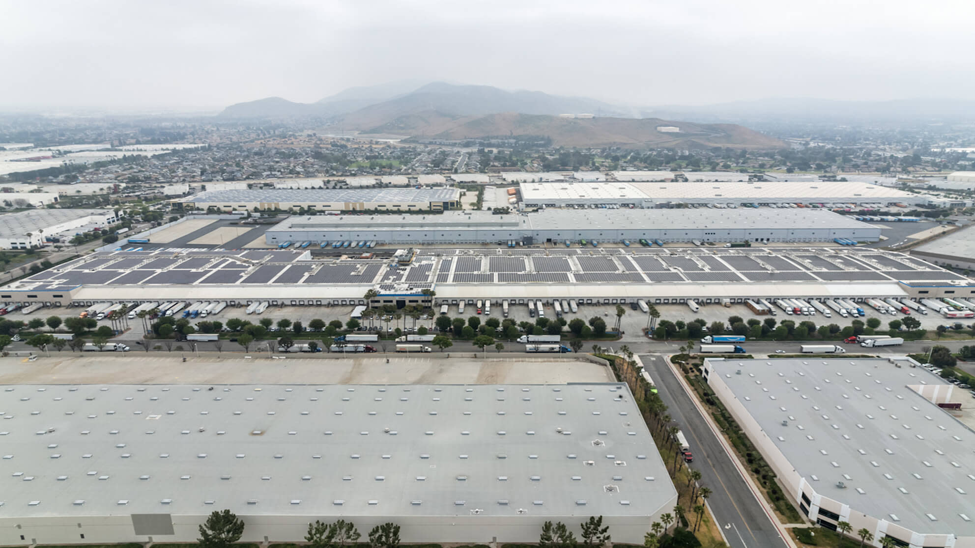 Aerial view of large industrial warehouse complex with solar panels and parking lots near mountains.