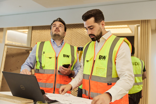 Two men in yellow safety vests reviewing plans and using a laptop indoors