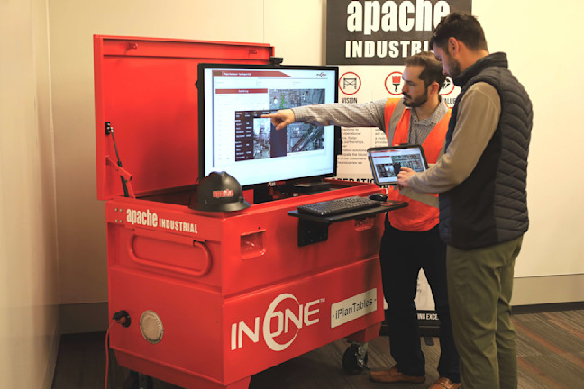 Two men review digital plans on a screen and tablet by a red industrial cart.