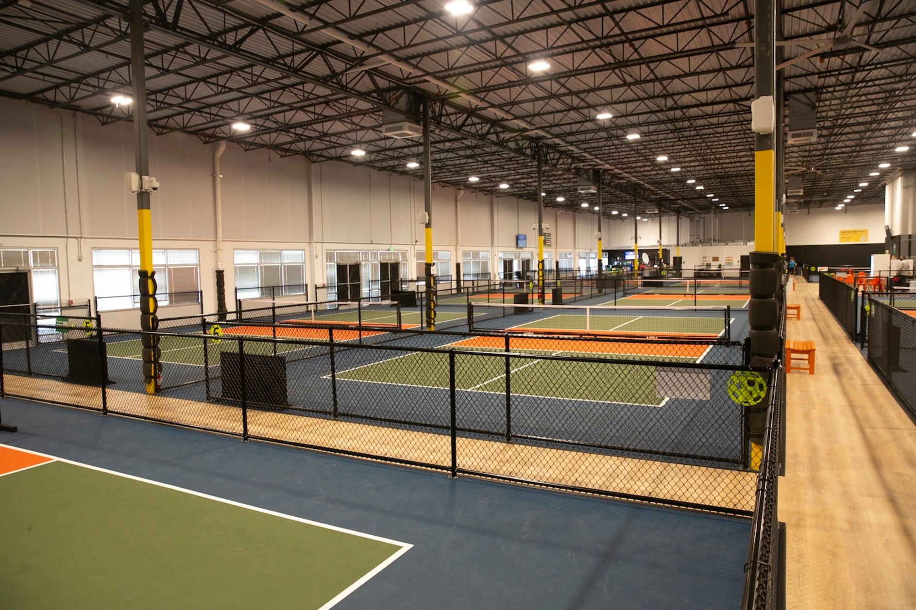 Indoor pickleball courts with orange and blue playing surfaces separated by black nets in a large warehouse facility.