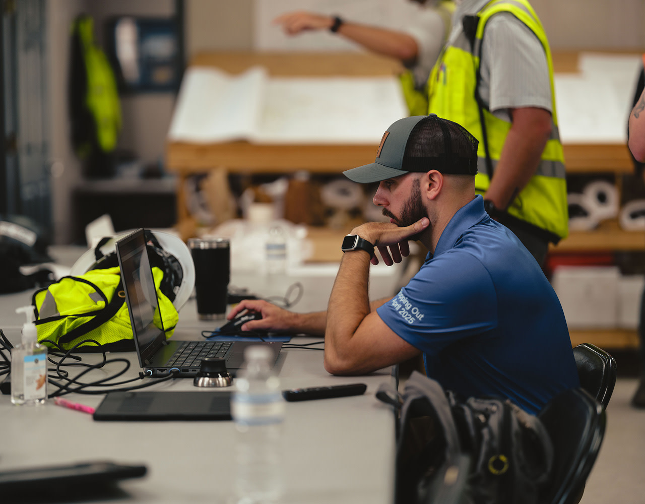 Man in blue shirt and cap working on laptop at a cluttered table.