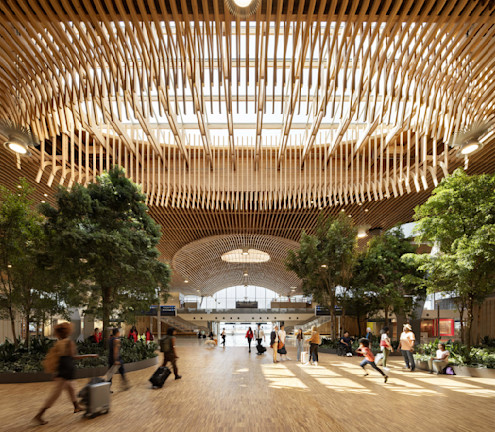 Spacious indoor area with wooden ceiling, trees, and people walking with luggage