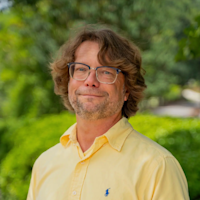 Man with brown wavy hair and glasses wearing a yellow button-up shirt outdoors with green foliage background.
