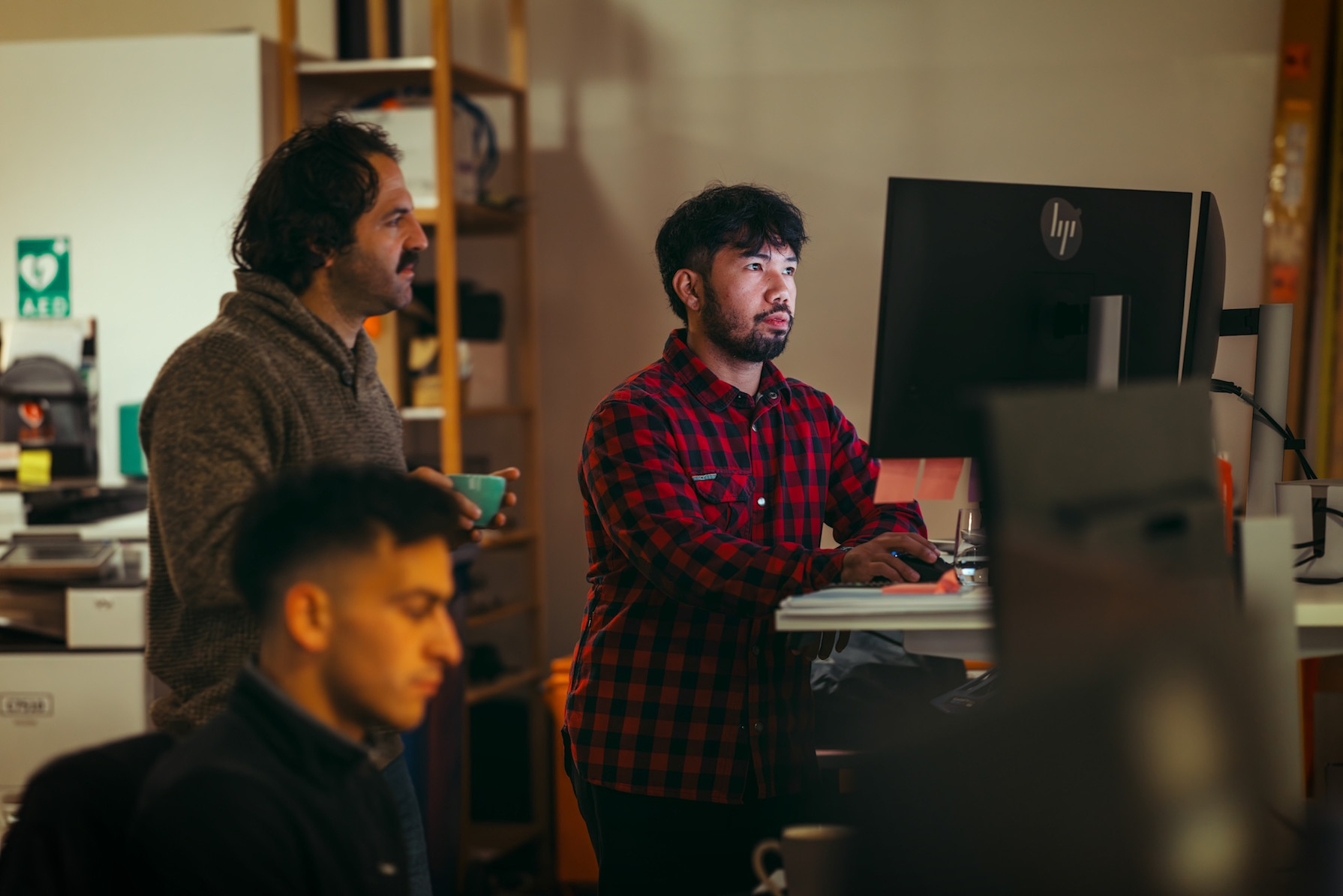 Three men collaborating at computer workstations in an office environment with warm lighting.