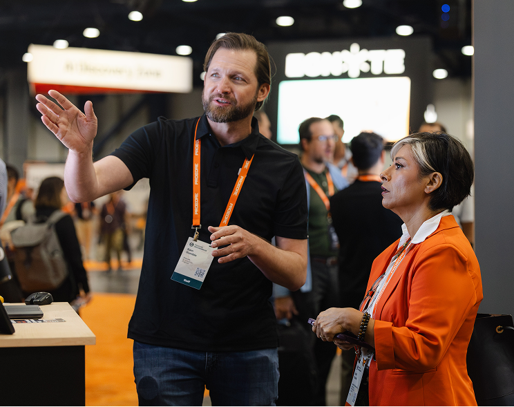 Man in black shirt with orange lanyard gestures while speaking to woman in orange blazer at conference event.